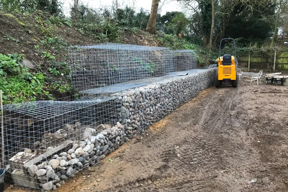 Gabion retaining wall under construction with stone fill, wire baskets and compact dumper