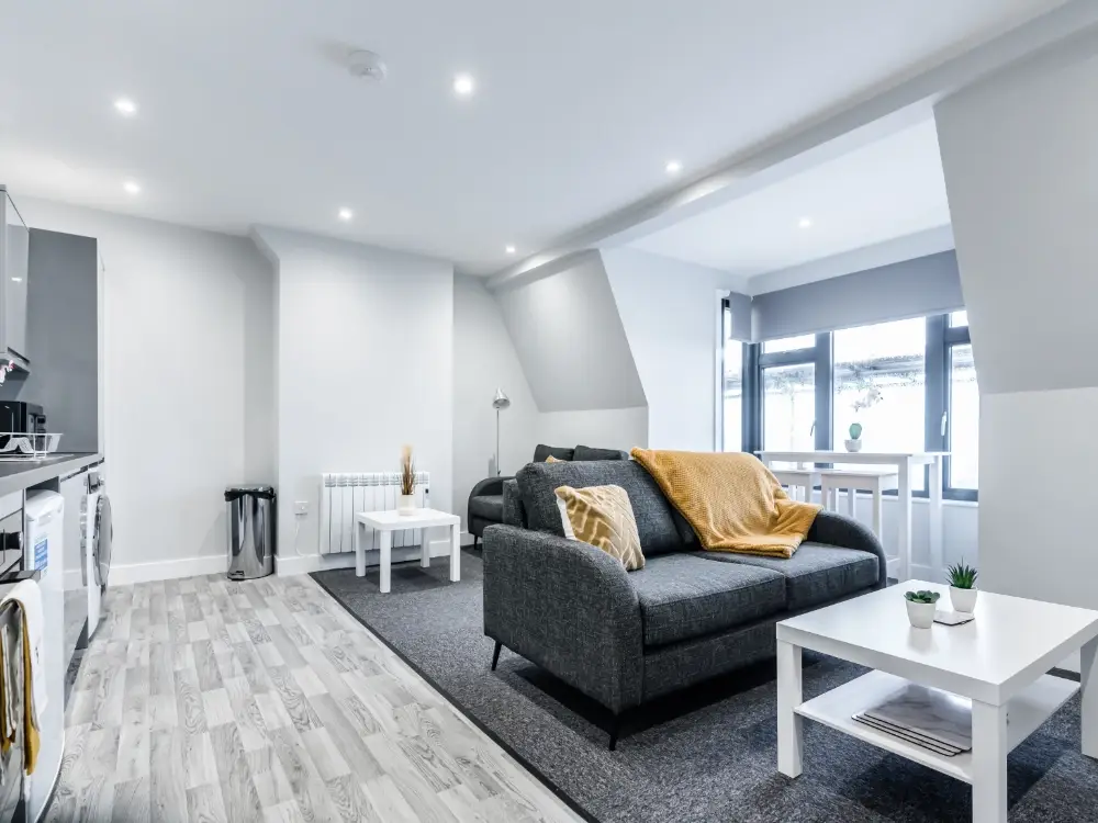 Modern open plan living room with grey sofa, yellow throw, white coffee table, grey carpet, and light wood flooring